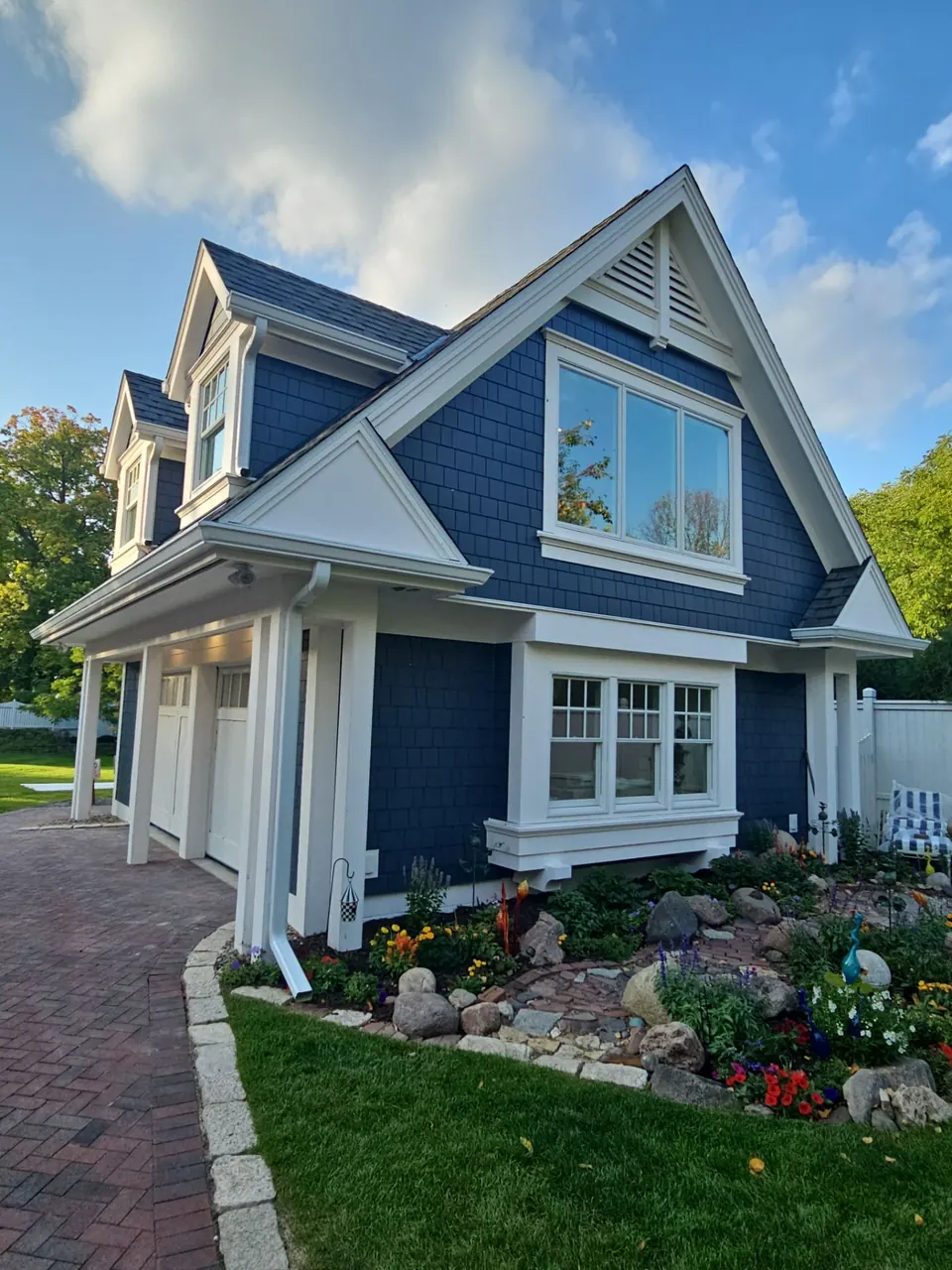 Navy shingle-sided home with white trim, a gable window and a dormer above the garage, with a paver driveway and rock garden in front.