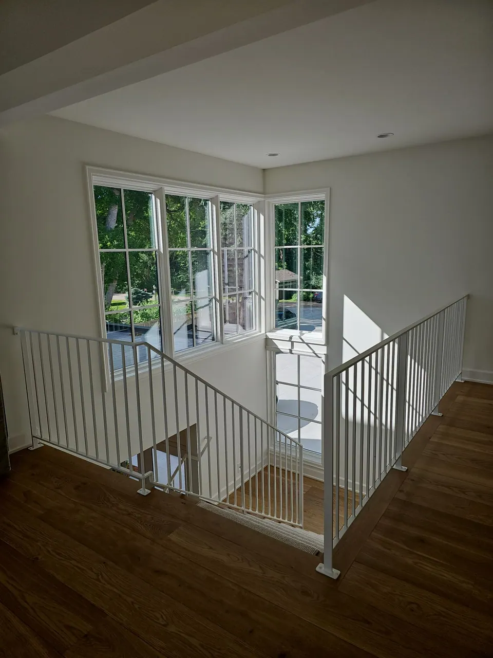 Empty upstairs landing with fresh white walls, a white metal railing, and a bank of corner windows over hardwood floors.